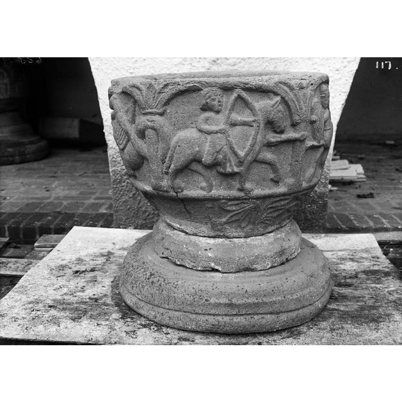 Source caption: "Dopfunt av sandsten, 1100- eller 1200-talet." [NB: the position of the font in this photograph should not be taken as indicative of its position in relation to the altar, as the font was being rotated for the photograph against the same background] -- there is another font here, partially visible in the background, inside the arcade, on the left