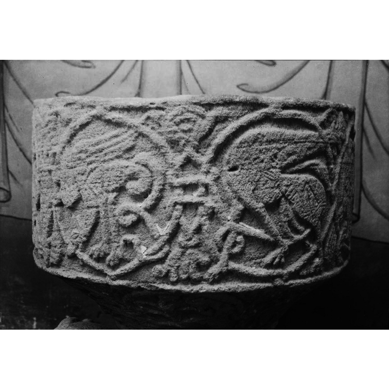 Winged creature - facing the left, legs straight and head bent down biting the frame. The frame has a life of its own - vegetation [seen here on the left side of the image -- NB: the original KMB image has been flipped horizontally to match the one in Sveriges kyrkor [cf. FontNotes]]