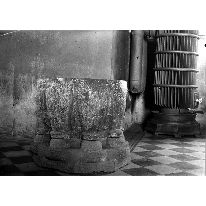 the medieval font already located in the new church that had been consecrated in 1902; the font and the heating stove seen here next to it were located in the southeast corner of the nave --  Source caption: "Suntak [...] Interiör [...] I nya kyrkan [...] 1918 Fotograf Roland, Anders"