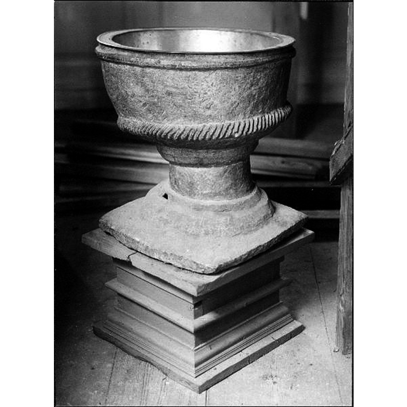 Source caption: "Älgarås kyrka [...] Dopfunt på gamla postamentet. Huggen i täljsten. Cylindrisk cuppa i nedre kanten prydd med repstav. Fot överst rund i flera avsatser, nedtill fyrsidig platta." -- the font, made of single block of soapstone, needs a tall plinth to raise it to a functional height