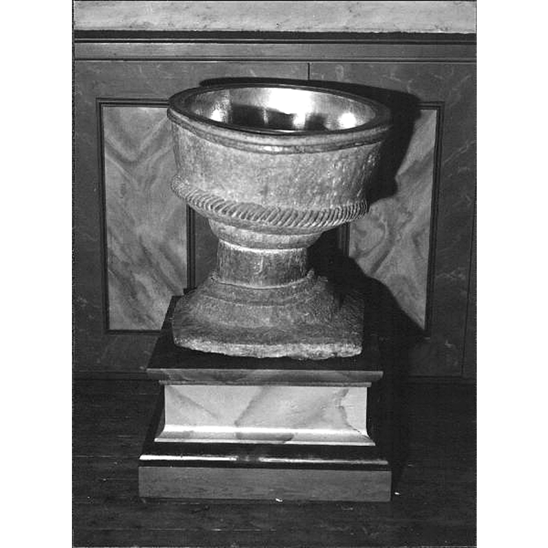 Source caption: "Älgarås kyrka [...] Dopfunt på gamla postamentet. Huggen i täljsten. Cylindrisk cuppa i nedre kanten prydd med repstav. Fot överst rund i flera avsatser, nedtill fyrsidig platta." -- the font, made of single block of soapstone, needs a tall plinth to raise it to a functional height; the irregularity of the work is clearly noticeable in this photograph