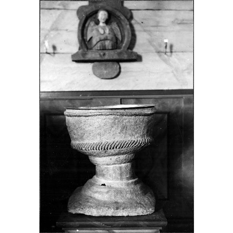Source caption: "Älgarås kyrka [...] Dopfunt på gamla postamentet. Huggen i täljsten. Cylindrisk cuppa i nedre kanten prydd med repstav. Fot överst rund i flera avsatser, nedtill fyrsidig platta." -- the irregularity of the work is clearly noticeable in this photograph