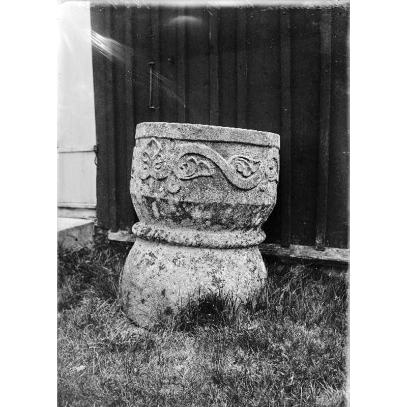 Source caption: "Ölsremma kyrka [...] Dopfunt av sandsten. Romansk ranka på cuppan, repstav om livet, foten oornerad." -- the medieval font is seen here ouside, by the west end of the porch [vapenhuset], obviously disused