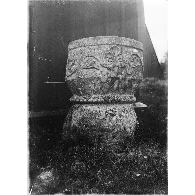 Source caption: "Ölsremma kyrka [...] Dopfunt av sandsten. Romansk ranka på cuppan, repstav om livet, foten oornerad." -- the medieval font is seen here ouside, by the west end of the porch [vapenhuset], obviously disused