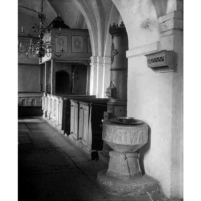 Source caption: "Hannas kyrka [...] Interiör mot nordväst." -- showing the medieval font against the north pillar of the chancel arch, near the old 'kakelugn'