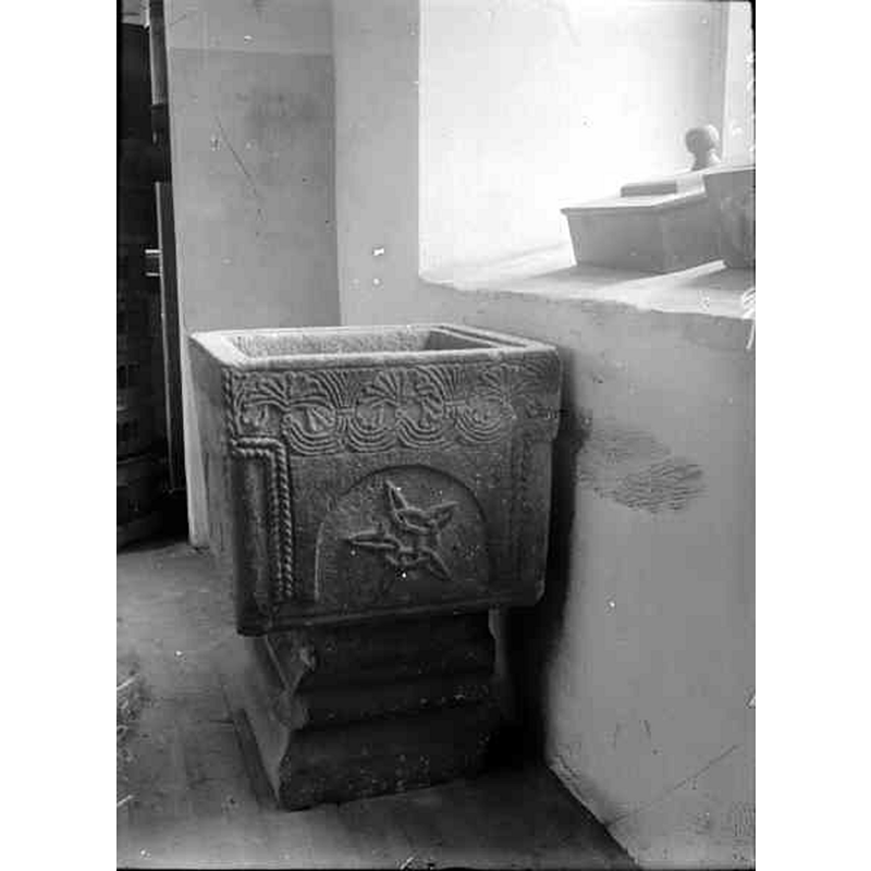 Source caption: "Nävlinge kyrka [...] Dopfunt av kalksten. Huggen palmettfris och rosetter. Spår av gul och röd färg." -- of the two objects on the window sill, the farthest appears to be the font cover