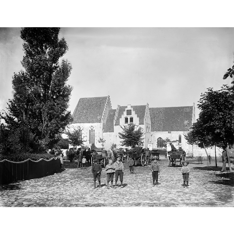 Source caption: "Skanör Church (St. Olof Church), Skåne, Sweden [...] Människor, hästar och vagnar utanför Skanörs kyrka (Sankt Olofs kyrka), ursprungligen byggd på 1200-talet. Kyrkan byggdes ut i etapper under 1300- och 1400-talet. / People, horses and carriages outside Skanör Church (Saint Olof Church) in Scania, originally built in the 13th century. The church expanded gradually during the 14th and 15th centuries."