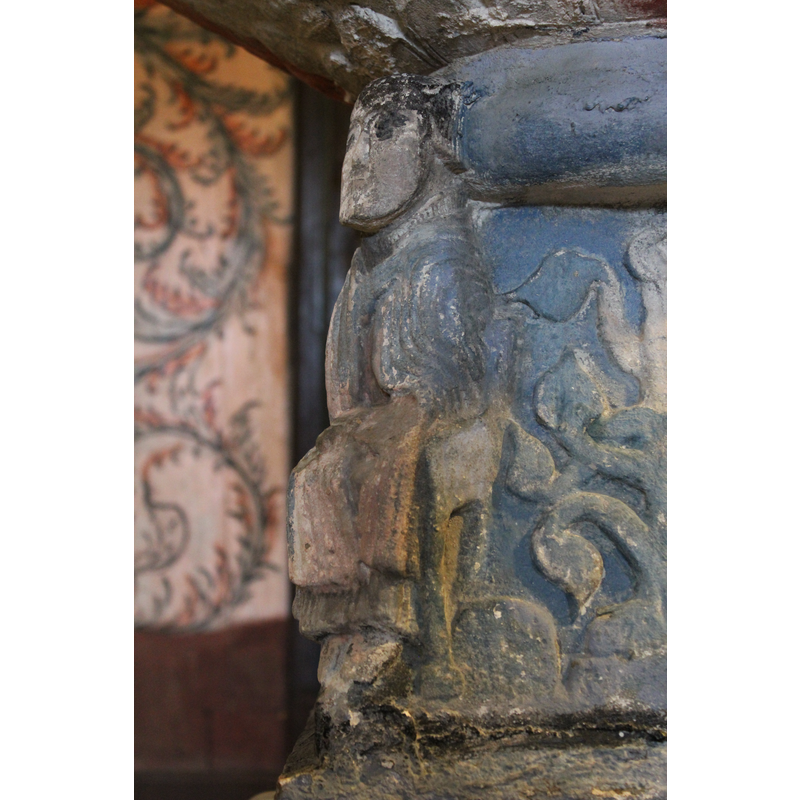 The first figure is a cleric, seated, full front, neat short hair, the right hand comes out of the mantle and holds the left side of the mantle near the neck. The right hand has been chipped off but appears to hold a book on his lap.