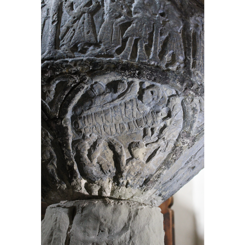 it this is a Nativity, it is a unusual one, and out of context with the rest of the programme on this font; the centre of the scene is occupied by an infant tightly wrapped in a cradle; above it, another supine figure, perhaps an infant as well; in the left-hand corner, however, two seated figures point [in typical Hegvaldian manner] to the cradle