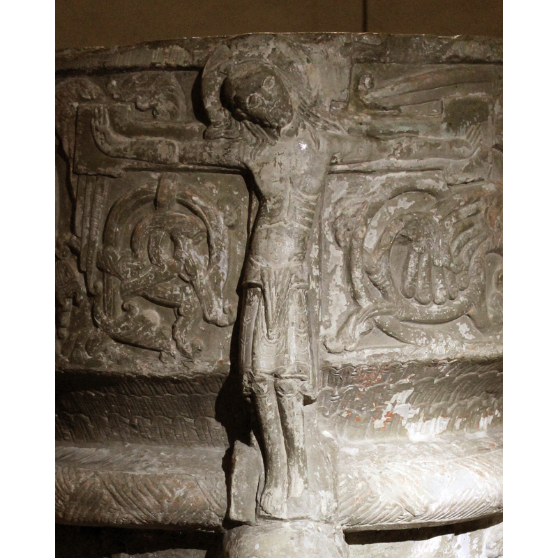 The Crucifixion of Christ is one large figure which wraps around the bowl. His arms and cross embrace the font. The cross is a cross pattee/potent (it has four square, boxed, ends). His hands point up, with thumbs lying inward on palm. His leans to his right shoulder. His legs and knees are slightly bent forming a s-shape on the cross. He has long hair falling over his shoulder, is bearded and haloed. His loincloth goes to the knees. There are detailed physical features such as torso, ribes and navel. Two circular, acanthus, spiral motifs are carved under the arms of the cross, on both sides. To the right of the Crucifixion is the symbol of the apostle St. John.