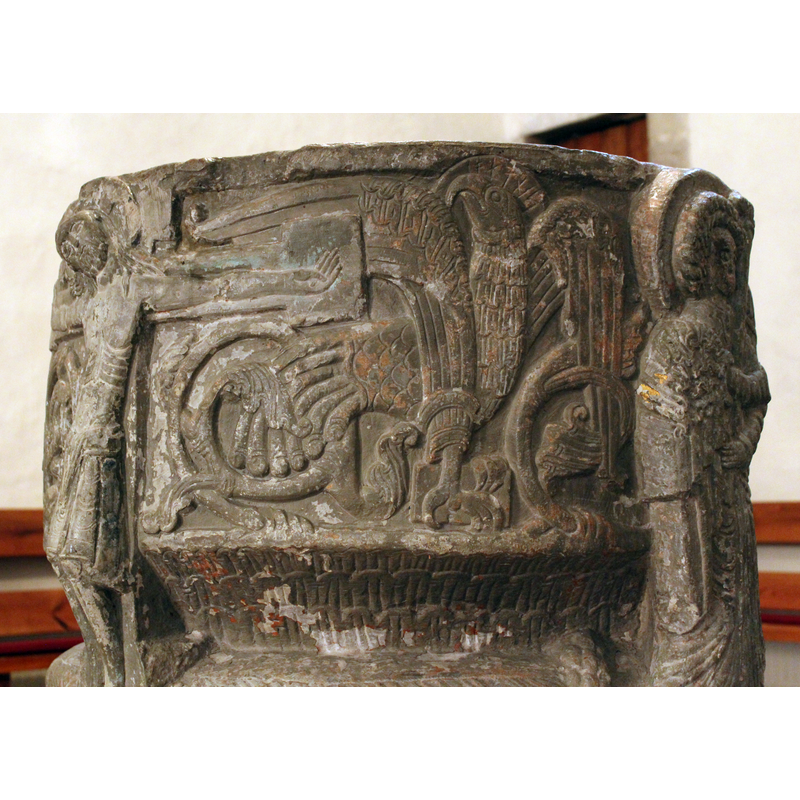 The eagle has its right wing raised and extended over the right arm of the cross. The haloed head is raised, looking back to the cross. Runic inscription in the halo. The left wing folds down at head height. Some of the acanthus vegetation evident under the arm of the cross continues to ornament and is entangled with the eagle's wing. The detailing of the beak and feathers is very fine. He holds a book in his claws.