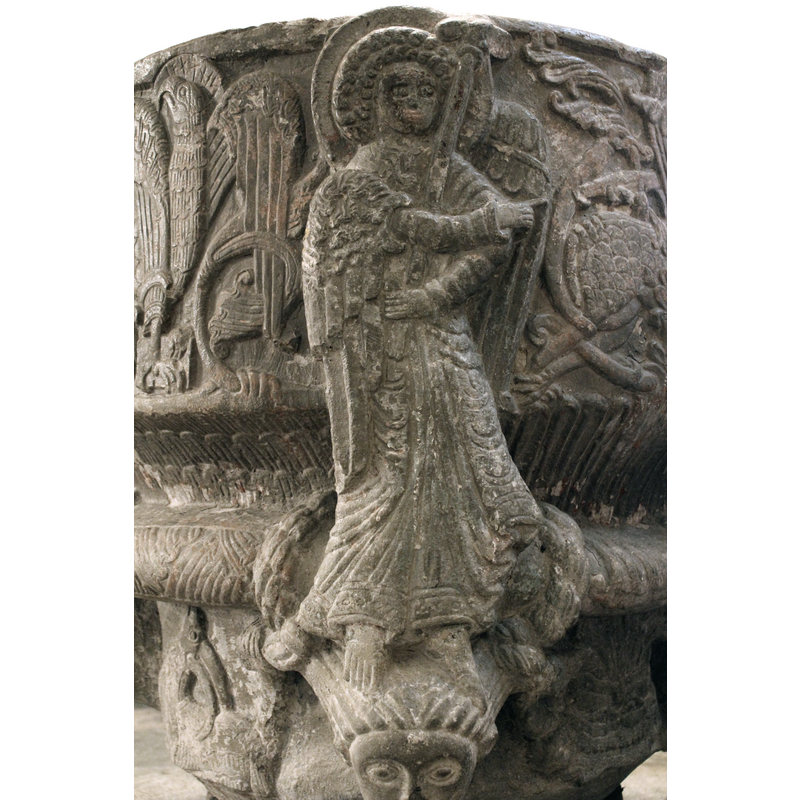 The figure of Gabriel is full-frontal, haloed with a large, curly head of hair in two rows. The wing to the right of the composition appears behind him. The wing to the left of the composition folds over his right shoulder and along the body. The body is slightly s-shaped with richly draped garments. His left arm is bent to the height of his midriff and holds a short staff which could have been a fleur-de-lis. His right arm is laid across his chest and points out with the index finger towards the right to the Virgin. Both feet are visible and stand on a female head with braids.