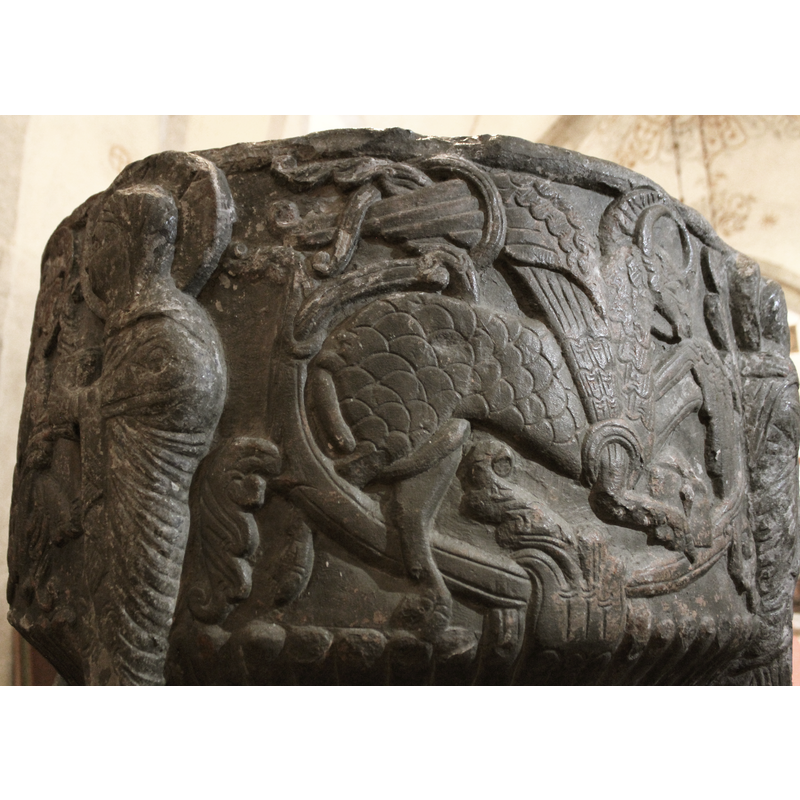 A winged ox which faces right. The head is haloed, with a Runic inscription in the halo. The front legs/hoofs hold a closed book and the scene has a background of vegetal ornamentation which is intertwined with the body, finely carved with many details.