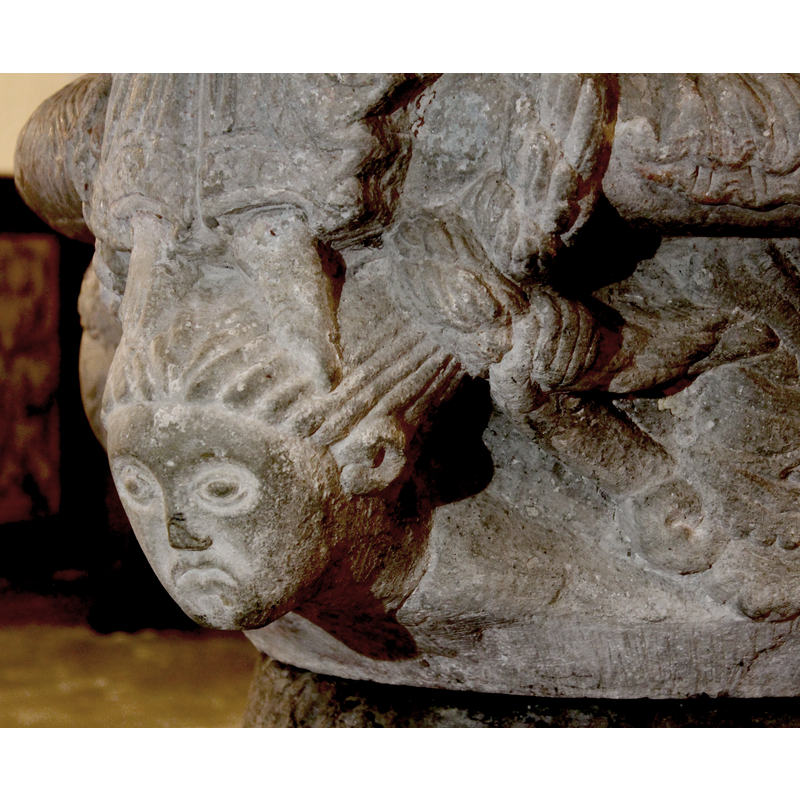 Head of woman with braids. Big wide eyes and mouth's sides are pointing down. Hair pull back tightly and the braids are wrapped around the centre ring and continue on the two left and right scenes. The braid is thick and serpent-like. [NB: the hand holding the braid on the right appears to belong to the inverted head on the right side of the base -- it may have been the artist's intention to create such an ambiguous image]