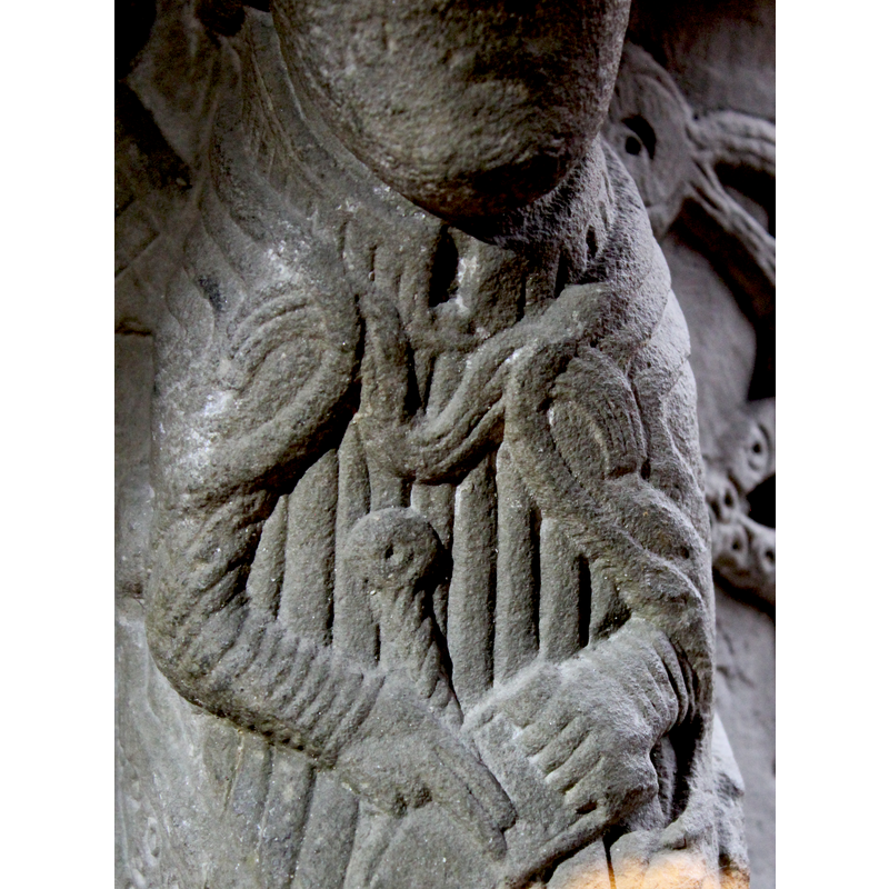 Untonsured head; the figure wears a rich cope [cappa pluvialis] tied with a clasp [morse] and embroidered pointed shoes; he holds a scroll with his left hand while two fingers [index and middle] of the right hand point to it; what appears to be the handle of a sword or dagger shows behind his hands