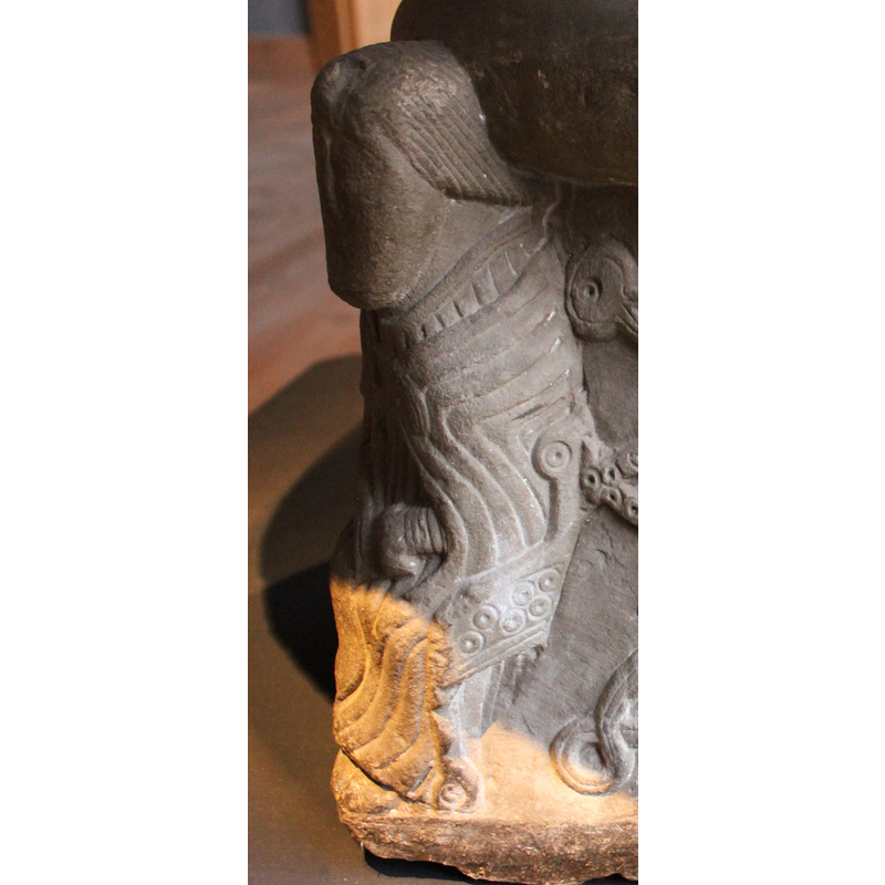 Untonsured head; the figure wears a rich cope [cappa pluvialis] tied with a clasp [morse] and embroidered pointed shoes; he holds a scroll with his left hand while two fingers [index and middle] of the right hand point to it; what appears to be the handle of a sword or dagger shows behind his hands