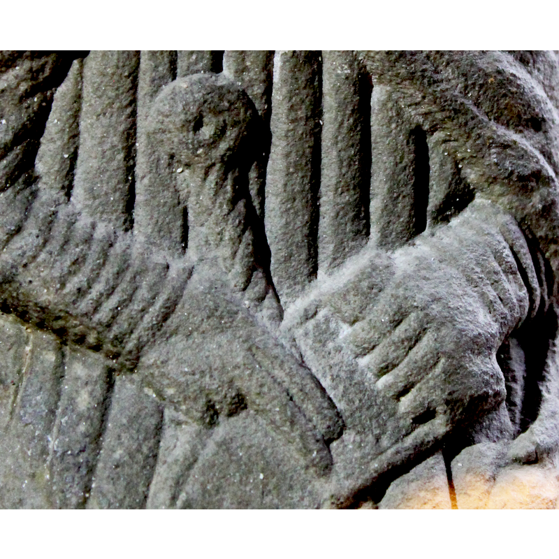 Untonsured head; the figure wears a rich cope [cappa pluvialis] tied with a clasp [morse] and embroidered pointed shoes; he holds a scroll with his left hand while two fingers [index and middle] of the right hand point to it; what appears to be the handle of a sword or dagger shows behind his hands