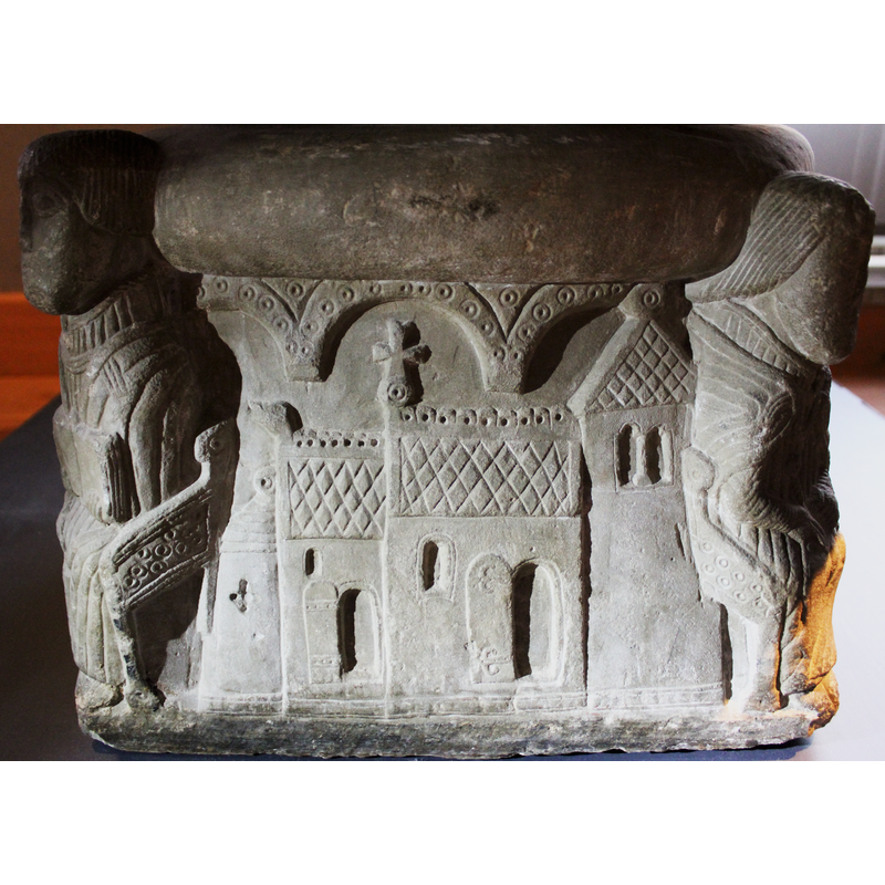 on the side between the seated tonsured monk and the seated cleric (?) holding a scroll -- North view of a Romanesque church building.of four elements: the east apse on the left, then [L->R] the chancel volume with the animal head on the top left end; next right is the volume of the nave topped with the cross; at the extreme right, the western tower. The two entrances on the north side are probably an error or liberty of the designer of the font, as such entrances [one to the chancel, another to the nave] normally occur on the south side in Gotland church buildings, not on the north side, where, if there is an entrance at all, is usually a single one. Notice the arcade of three round arch-heads decorated with beaded-tape pattern above the church building.