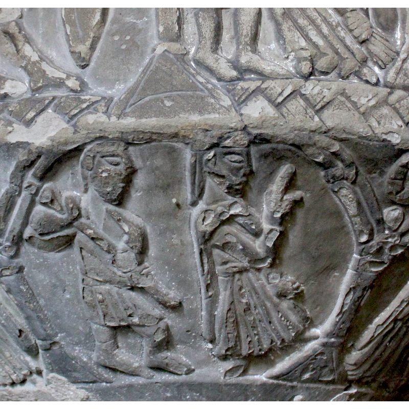One arch - scene #5 - on underside. Two figures, both sitting, the figure on the left, full frontal, holds a sword in the left hand leaning on his left knee, pointing upward. The right hand is visible at right side of the chest - oath posture (?). The head is turned to the right in profile. No head covering visible, worn. The figure on the right, the upper body leans to the left, head facing left, wears a crown, 3 points, the right arm is bent at the elbow and raised diagonally across the composition, index finger extended. It has six fingers on the hand and the cuffs are very wide - but this does not appear to be a woman. The left arm holds a sword pointing upward, leaning on his left knee. The spandrel holds a four-pointed star or flower, not a rosette like the other spandrels.