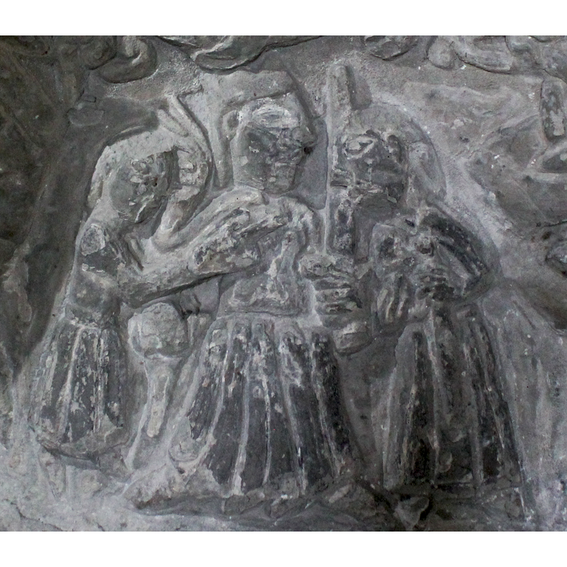 Three figures. The centre one is enthroned, sitting full frontal, the head is crowned with 3-pointed crown, looks to the right. The right arm is raised at shoulder, the hand has two fingers outward pointing to the left, diagonally up. The left arm holds a sword pointing upward. The figure on the left stands in profile, the right arm points diagonally, with an extended index finger to the figure on the extreme right. This figure, on the extreme right, faces the left, the king, the right arm appears to be holding an object close to its body, the left arm is bent at the elbow and holds what appears to be a sword.