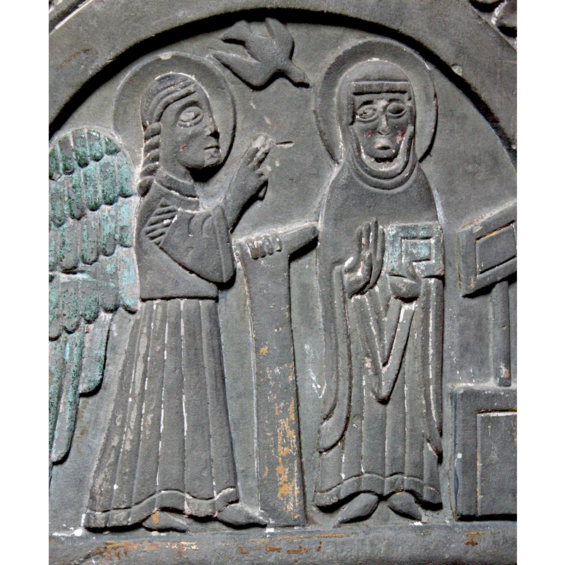Gabriel, in profile, on the left, standing and resting left hand on the lectern [or scroll] and his right hand is raised finger pointing to upward to the Holy Ghost (dove)…the lectern might be a scroll (?). Mary wears a cowl and stands (full frontal) with right hand raised showing inside of her palm and left hand holding a book. To her right stands a lectern. Dove above, between the two haloed figures