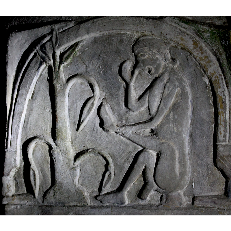 on the southeast side of the basin [orientation is approximate] -- a figure to the right sits on a little pedestal. His left hand holds his left leg by the ankle. He is naked. His right leg rests on the ground. His right hand is raised to his face. Has a large penis. To his left is a bush or tree with branches.
