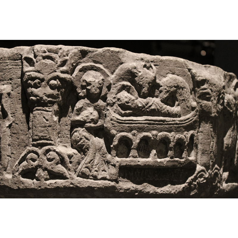 This scene is carved in two arches, both now incomplete. The arch to the right contains, a bed, the headboard of which is a lion, facing the left; Mary lies in bed, covered head in profile, haloed; her right arm is visible over the covers, hand resting on her abdomen; she wears very wide sleeves. The bedcover is long and richly adorned. There is a row of hatched lines inscribed on the lower edge of the bed. The top right hand corner of the scene is missing, but a hand appears to be coming from it [NB: do other images by this artisan have a similar image?]  In the second arch of this scene, there is a large boat-shaped raised on an  arcaded bottom [cf. English Nativity scenes], six columns and five arches [cf. other example]. The child is swaddled, haloed head to the left, cross in haloe, and the head looks at the viewer; above are the two heads of ass, to the right, and the ox, to the lef). Further to the left sits Joseph on a rock or stool, his legs in profile covered with a long gown; his upper body is full frontal, haloed, long hair, both hands hold an object to the middle, round or oval shaped. The upper part of this scene is also missing.