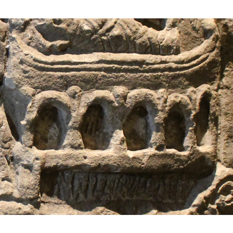 a very sophisticated manger shaped like a boat, raised on a deeply-carved arcade of five round arches with ball motif in the spandrels; the space below is incised with hatched lines [not an inscription]