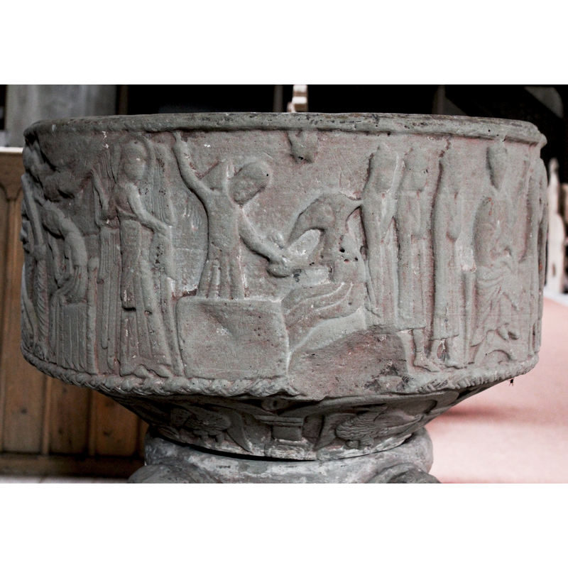 on the east side of the basin [orientation is approximate] -- at the left an angel in profile, but showing wings, pointing upward with its right hand to the star behind; the left arm has fingers in benediction. The figure in the middle standing behind a rectangular object, (altar, table) in 2/3 pose, right hand raised up and pointing upward, head slightly bent down toward the right; he wears a hooded garment often used in the depiction of shepherds on fonts of the period; his left arm is holding the right hand of the seated figure to the right. The figure to the right is a seated old man wearing a long beard and a hood. A star above the two shepherds.