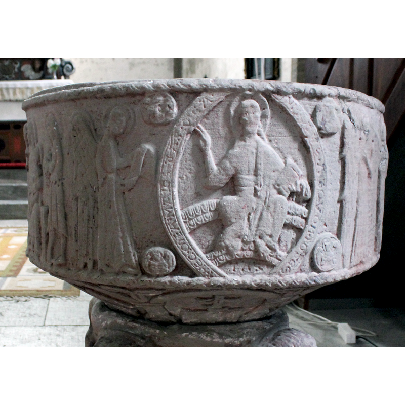 on the west side of the basin [orientation is approximate] -- a haloed angel stands in profile at the extreme left, both hands holding either a scroll or a stole. Christ sits on a rainbow inside a mandorla framed by four medallions in which are the symbols of the four Evangelists; the mandorla bears an inscription [cf.InscriptionArea]. Christ's throne is a two-tiered curved bench and his feet rest on a similar structure. He wears a crossed halo; his right hand is raised in benediction; his left hand holds a book and part of his robe; his feet are bare.