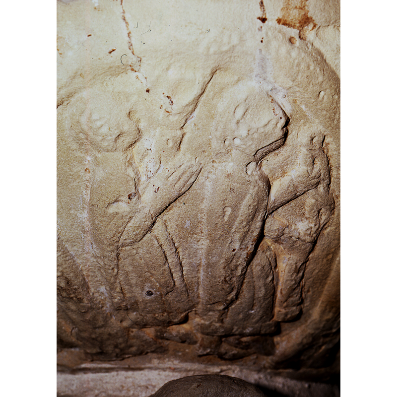 Two figures standing in profile, facing right, wearing mantles; the figure to the left, has arm bent at elbow, 45* angle, palm extended; the left hand is raised or holding up an object in the background? Torch? Or just the hand? The figure on the right, the left arm holds an object at the middle (long object); the lower part of the object appears cylindrical and the upper part is twisted or braided and is the same length as the other one; it is being held by the figure's right hand. These two figures may be related to the next two in the arch to the right