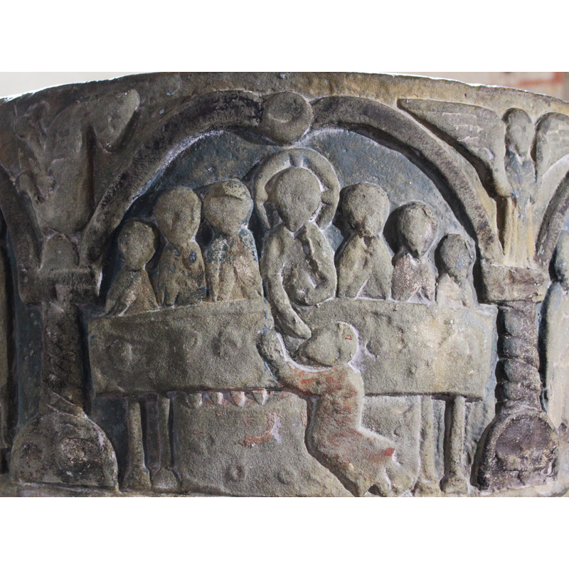 At the centre of the scene is a large rectangular table on four legs. At the centre of the table is Christ, full frontal, cross-haloed, holding in his left hand an object to his middle - the right hand reaches across the table to the viewer's side, where a figure is kneeling with arms raised and being fed by the hand of Christ. To each side of Christ are three figures facing the front; six in total. The figure to Christ's right has his hand raised diagonally across his chest with his finger pointing to Christ. In the spandrel is an angel with open wings, and hands raised same as before.