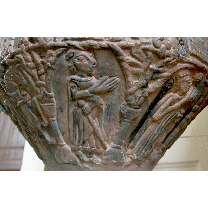 Male figure stands 3/4 profile, frontal. Dressed in long robes and cape. The right hand, palm extended, raised across his chest at 45* angle. The left hand fold intowards his middle and holds a staff with a knob. He wears either a hat or maybe a crown