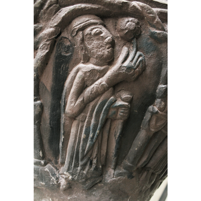 Figure in profile, dressed inlong robes and mantel and has ahead cover. The face is raised up and looking at the star. His right arm is bent at the elbow and the hand with finger extended touches the star. (Wiseman). The left hand protrudes from mantel and holds a knobed staff.