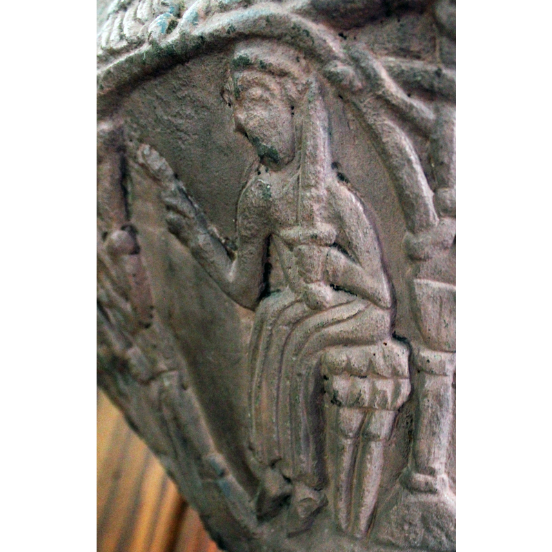 Figure of a man profile (3/4) sitting on bench. His left arm bent at elbow and holds a sword and points to the scene above with the two women. The right hand points up.