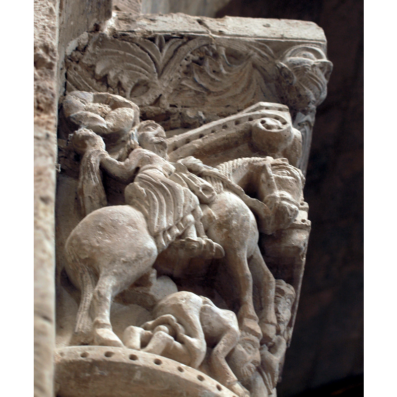 capital on the north side of the chancel arch: on the left side of the capital is a mounted male holding an object and/with a flowing cloth in the right hand; the left hand hold the bridle and a ring, or the ring is secured to the saddle; the horse tramples over a fallen bearded man who appears unarmed; the front side of the capital has a balcony with two males in it looking out; below the balcony a male figure leans on a short pillar (to which he may be tied?); the right side of the capital has a bearded man loading a stone into his sling, by the side of a mounted knight armed with a lance and a shield, and wearing a helmet; behind him is an unidentified beast
