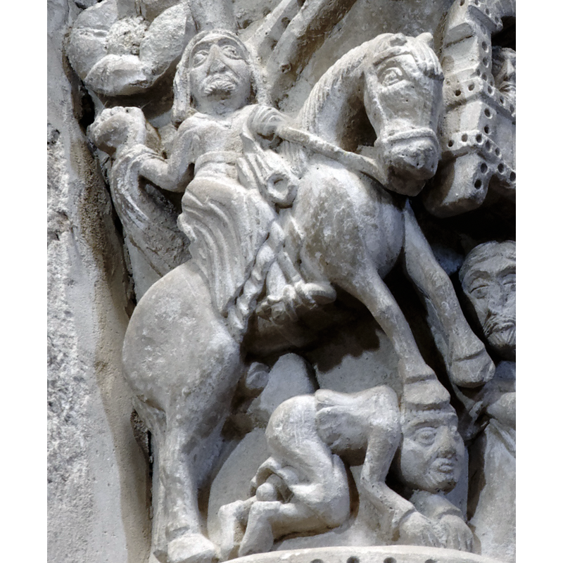 capital on the north side of the chancel arch: on the left side of the capital is a mounted male holding an object and/with a flowing cloth in the right hand; the left hand hold the bridle and a ring, or the ring is secured to the saddle; the horse tramples over a fallen bearded man who appears unarmed; the front side of the capital has a balcony with two males in it looking out; below the balcony a male figure leans on a short pillar (to which he may be tied?); the right side of the capital has a bearded man loading a stone into his sling, by the side of a mounted knight armed with a lance and a shield, and wearing a helmet; behind him is an unidentified beast