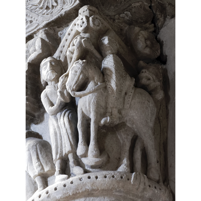 capital on the north side of the chancel arch: on the left side of the capital is a mounted male holding an object and/with a flowing cloth in the right hand; the left hand hold the bridle and a ring, or the ring is secured to the saddle; the horse tramples over a fallen bearded man who appears unarmed; the front side of the capital has a balcony with two males in it looking out; below the balcony a male figure leans on a short pillar (to which he may be tied?); the right side of the capital has a bearded man loading a stone into his sling, by the side of a mounted knight armed with a lance and a shield, and wearing a helmet; behind him is an unidentified beast