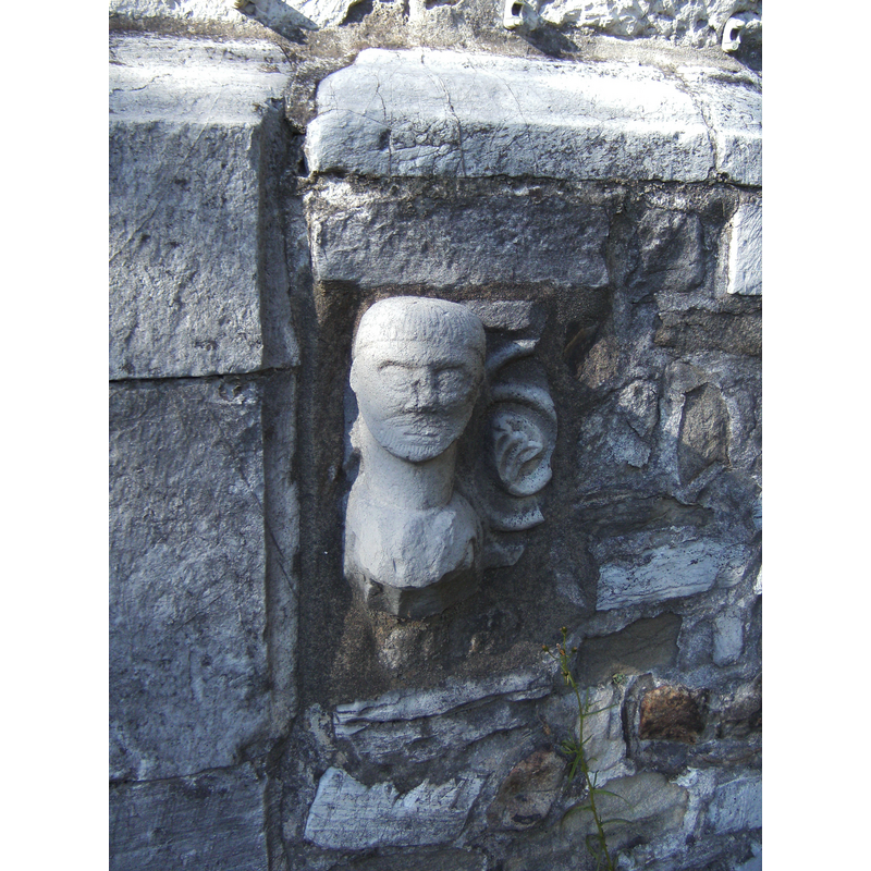 fragment of the font re-built into the exterior south wall -- Photo caption: "Fichée dans le mur extérieur du bas-côté sud, cette tête sculptée provient des fonts baptismaux du XIIIe siècle, aujourd'hui perdus."