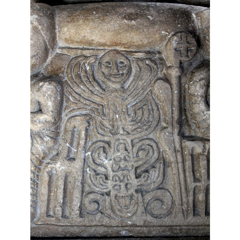 the upper part has a human figure with a large smiling head and branch-like upper limbs; the lower part appears to be a combination of two masks with scroll-like protrusions and wings -- on the south side of the base [NB: orientation is approximate]