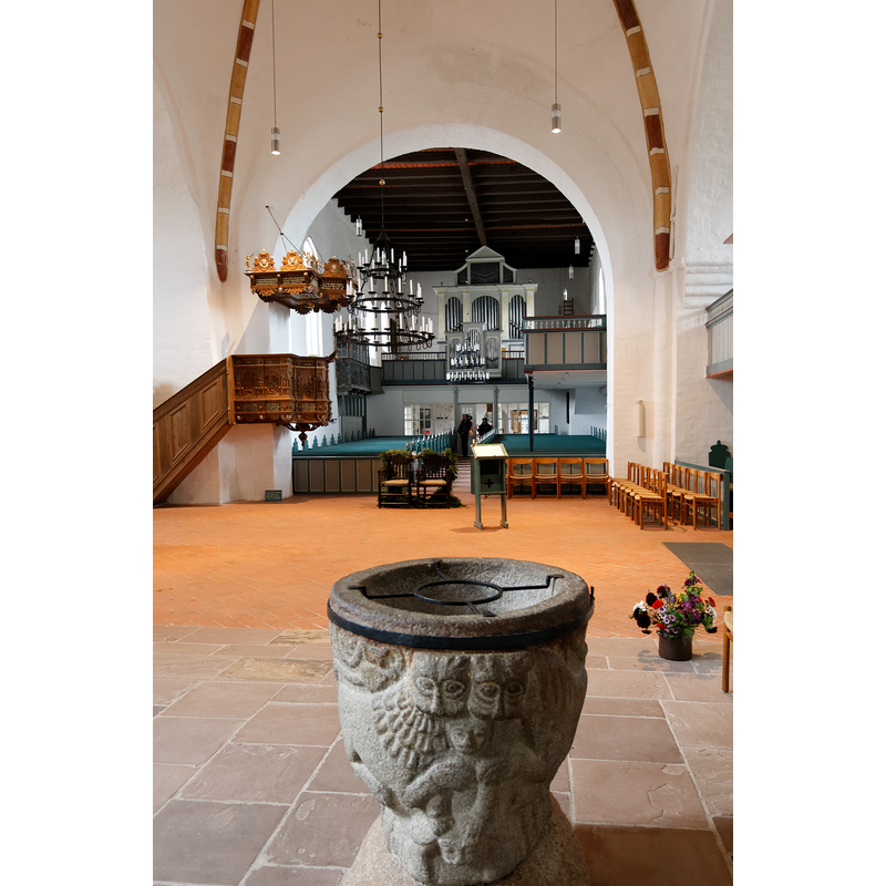 the old font, east side facing the camera, in the foreground -- Photo caption: "Blick durch das Mittelschiff auf den Altar / Mittelschiff der St. Johannis-Kirche in Nieblum auf Föhr mit Blick auf den Altar"