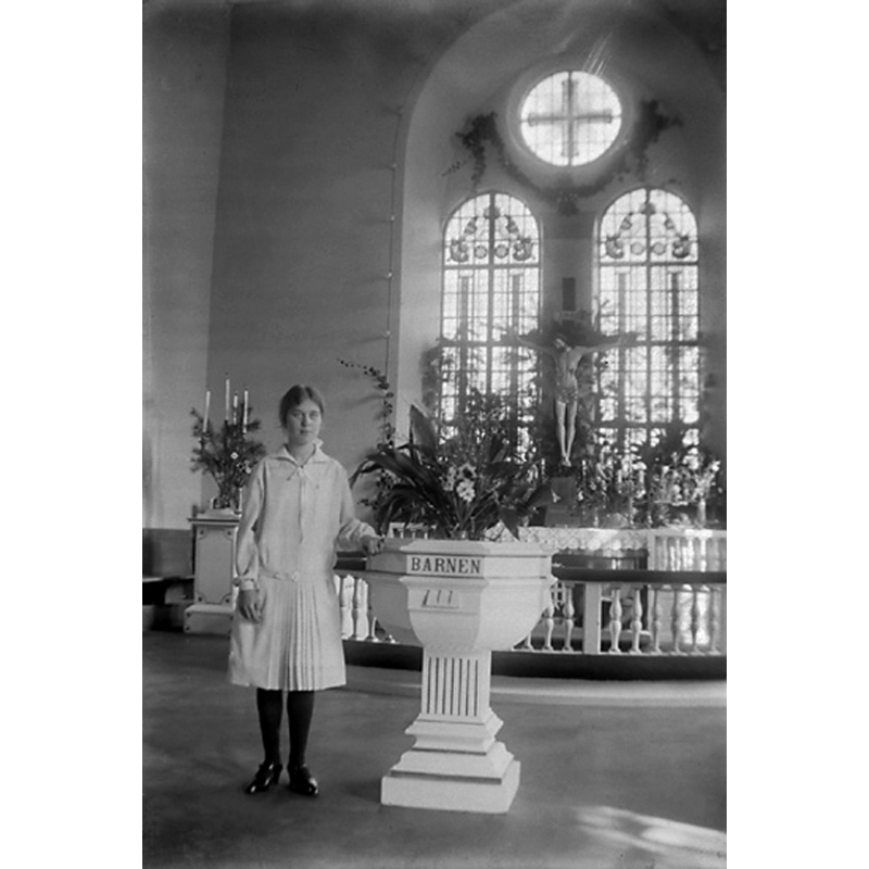 The Bohusläns museum source notes that the person posing next to the font is Gertrud Bonander at the time of her confirmation in 1928; the photographer was her sister