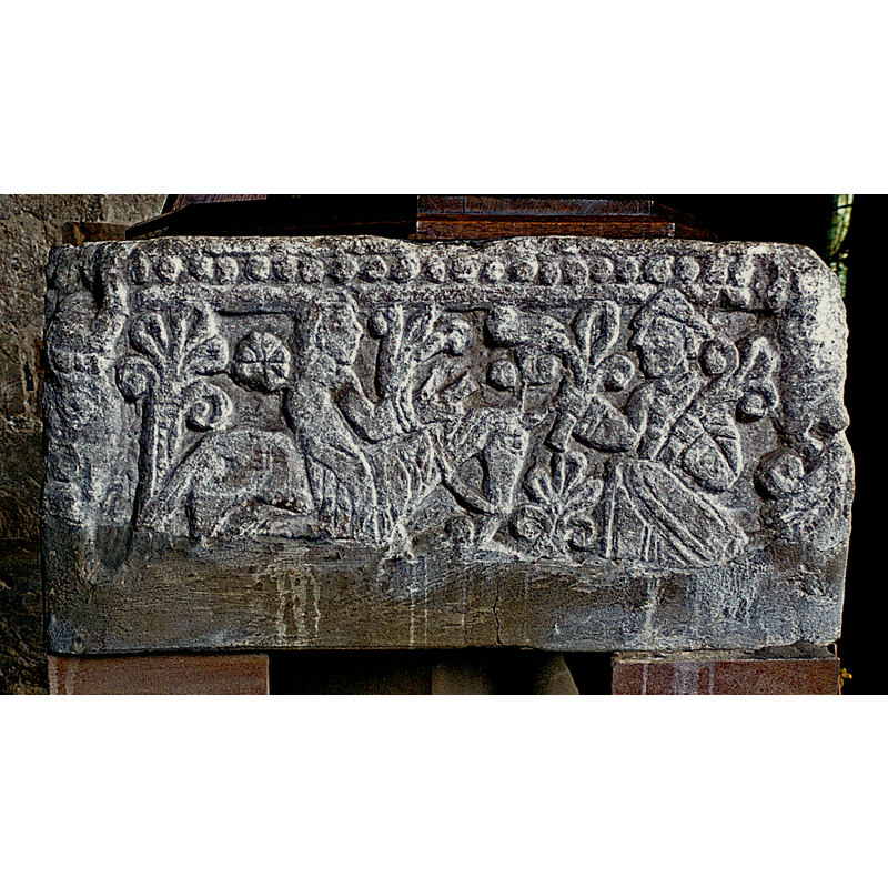 west side of the basin: a figure on horseback riding towards the right holds up a palm; to the right, a figure facing left is holding up palms; above is the row of large beads thta decorates all side of this font; on the right and left angles are deformed heads with vegetation stemming from their mouths