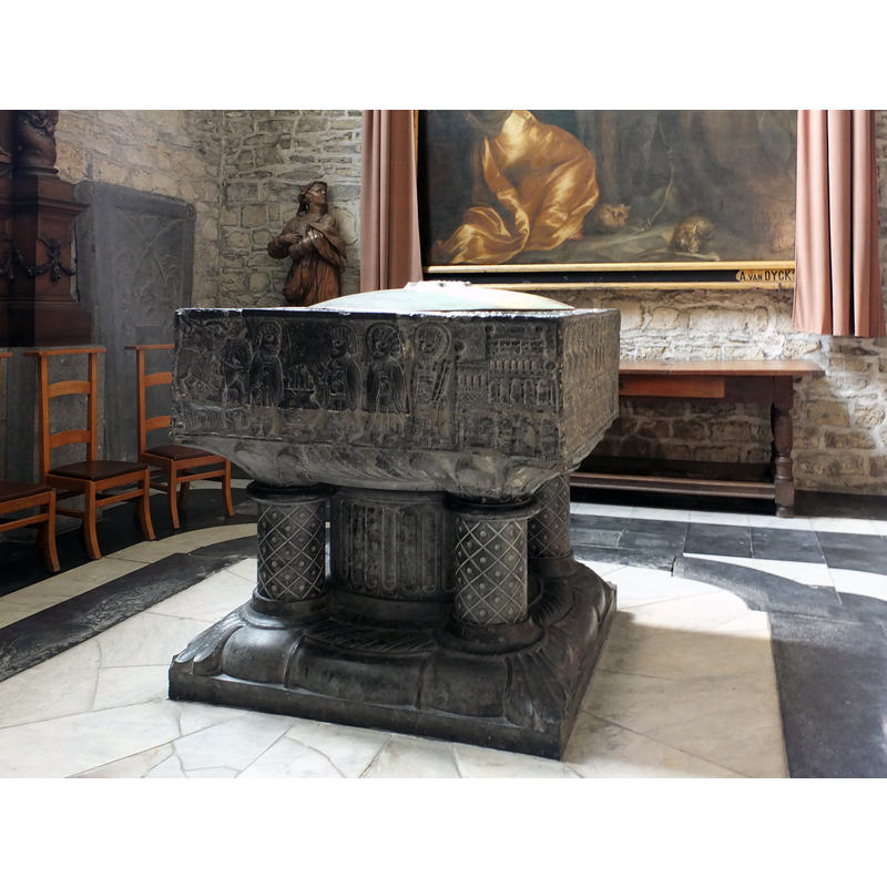 Source caption: "De elfde eeuwse Romaanse doopvont in Doornikse kalksteen in Onze-Lieve-Vrouwekerk van Dendermonde is het oudste kunstwerk van de kerk. Op de zijvlakken staan symbolische voorstellingen in verband met het doopsel. Twee friezen stellen de belangrijkste momenten voor uit het leven van de apostel Paulus: zijn bekering en zijn opname in de gemeenschap van de apostelen, gerestaureerd in 1858-1860 door J.B. De Pauw (Dendermonde) naar plan van architect L. Roelandt (Gent) met toevoeging van voetstuk; deksel met beeldengroep "doopsel van Christus" van 1862, hout en koper; witte marmeren wijwatervaten door beeldhouwer Walter Pompe (Antwerpen), 1774."
