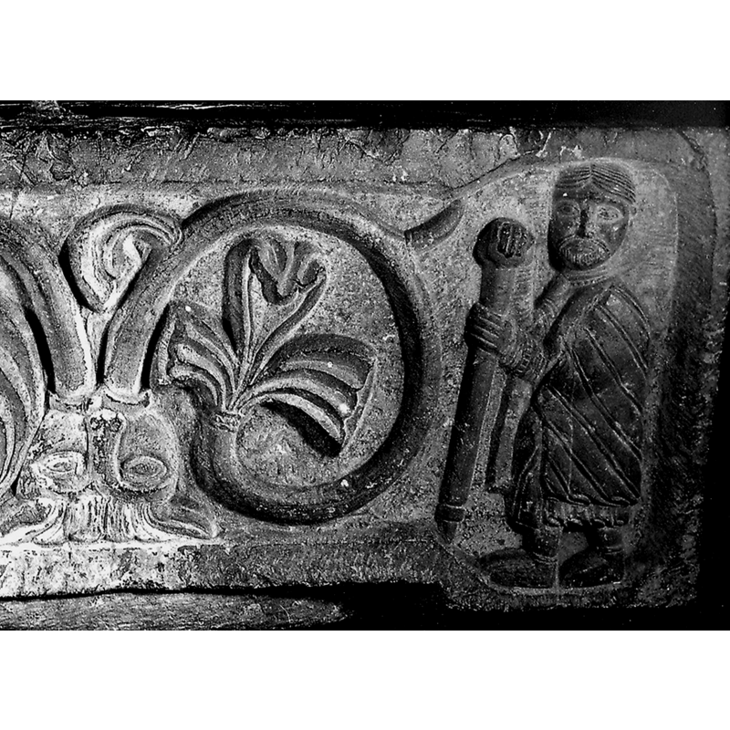 basin side 1:a figure in rich garments looking at the viewer and holding a thick adorned staff, at the right end of the side