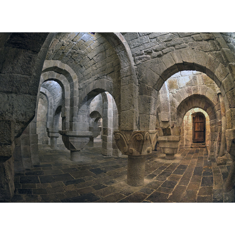 Source caption: "Esta cripta es un extraño espacio destinado, parece ser, a soportar el peso de la estructura de la iglesia que tiene encima. Este montaje de cortas columnas con grandes capiteles, irregulares y diferentes, está datado en la primera mitad del siglo XI."