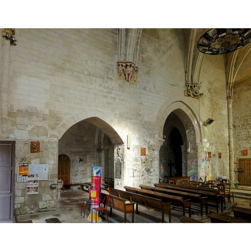 the font is partially visible on the left, just right of the wall-mounted stoup, left of the colourful stand -- the plan of the church interior has been much altered through the centuries -- Source caption: "Chœur, côté ouest : arcades ouvrant dans le croisillon sud et la croisée du transept. Le chœur étant désaxé par rapport à l'ancienne nef et la croisée, le croisillon nord ne communique pas avec lui."