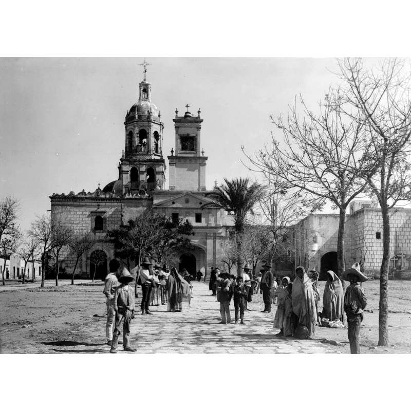 Source caption: "Photograph of the church of La Cruz, Queretaro, Mexico, ca.1905-1910. The open ornate dome on top and bell tower are prominent. A group of a couple dozen people are standing about the paved walkway which is sparsely lined with trees, leading through the plaza toward the church."