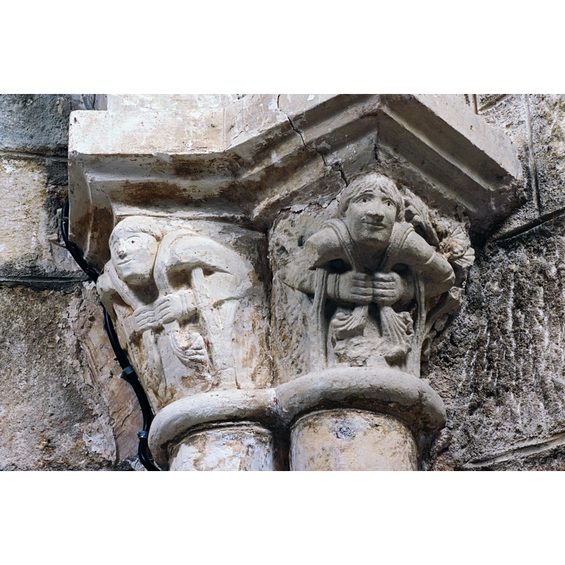 Source caption: "Le décor sculpté se compose de chapiteaux, en place ou déposés dans la chapelle nord. Outre les motifs végétaux, le décor comporte des personnages aux bras entrelacés se tirant la barbe et d’autres, aux angles de la corbeille, paraissent supporter de leurs bras le poids de l’architecture."