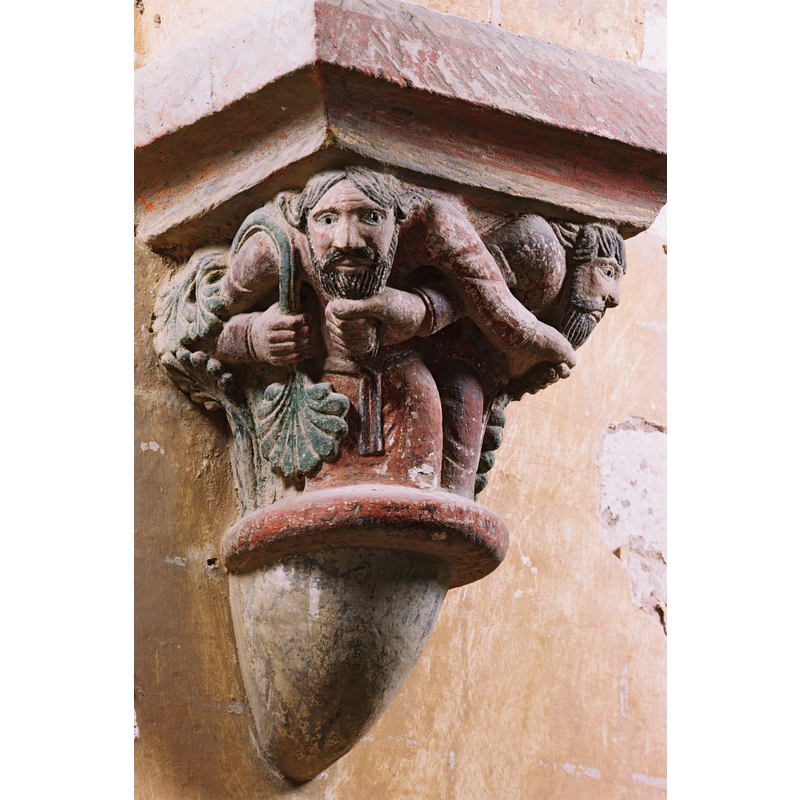 Source caption: "Le décor sculpté se compose de chapiteaux, en place ou déposés dans la chapelle nord. Outre les motifs végétaux, le décor comporte des personnages aux bras entrelacés se tirant la barbe et d’autres, aux angles de la corbeille, paraissent supporter de leurs bras le poids de l’architecture."